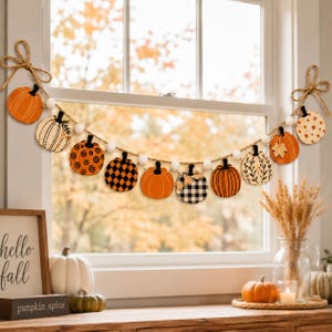 May include: A decorative garland featuring eleven wooden pumpkin cutouts in various orange, black, and white patterns, strung on a jute rope with white felt balls. The ends of the rope are tied into bows, showcasing pumpkins with stripes, checks, and dots.