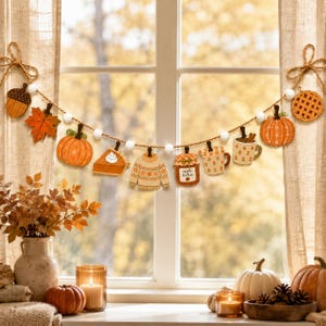 May include: A decorative garland featuring wooden cutouts of an acorn, maple leaf, pumpkin, slice of pie, sweater, jar labeled apple butter, mug, and a lattice pie crust, all strung on a rope with white felt balls between each seasonal shape.