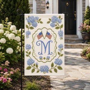 May include: A rectangular garden flag featuring a white background with a central scalloped frame enclosing a large blue letter M and two small American flags. The design is surrounded by blue hydrangea flower clusters and green leaves.