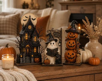 Puede incluir: Un conjunto de tres decoraciones de Halloween que incluye una casa encantada oscura e iluminada con luna llena, un fantasma con sombrero de bruja leyendo un libro de hechizos junto a un gato negro y tres calabazas apiladas con una escoba.