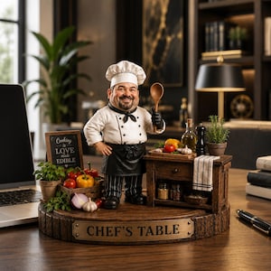 Op de afbeelding: Een kokfiguur staat op een houten voet met het opschrift CHEF'S TABLE, gekleed in een wit jasje, zwart schort en koksmuts. De kok houdt een houten lepel vast. Daarnaast staan een kleine houten tafel en een bordje met Cooking is LOVE made EDIBLE.
