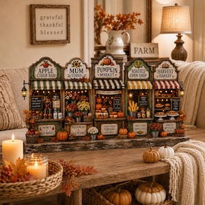 Könnte beinhalten: Eine dekorative Tischdekoration mit fünf Miniatur-Herbstläden: Apple Cider Bar, Mum Stand, Pumpkin Market, Roasted Corn Shop und Harvest Bakery. Jeder Laden verfügt über gestreifte Markisen, beleuchtete Innenräume und detaillierte Schilder.