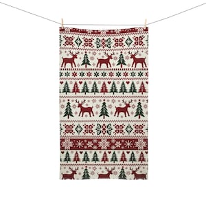 May include: A cream-colored Christmas-themed towel with a repeating pattern of reindeer, Christmas trees, snowflakes, and geometric shapes in red and green. The towel is hanging from a clothesline with a wooden clothespin.
