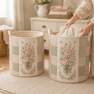 May include: Two cylindrical fabric storage baskets featuring a quilted patchwork design in soft shades of pink, green, and cream. Each basket displays a central panel with a detailed floral bouquet print, small butterflies, and side handles.