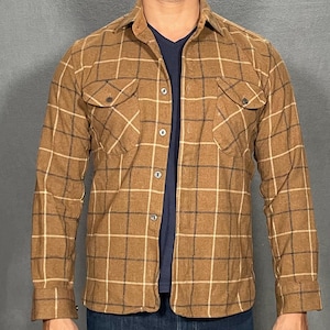 May include: A brown plaid shirt with a classic collar and two chest pockets. The shirt is worn over a navy blue v-neck. The plaid pattern features tan and dark brown lines. The shirt is made of a textured fabric.