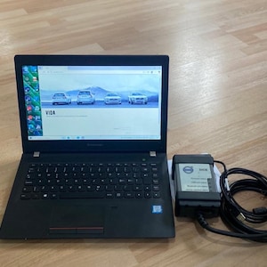 May include: A black Lenovo laptop with the VIDA software interface displayed on the screen. A black diagnostic tool labeled "DICE" is connected to the laptop via a black cable. The laptop is open on a light wood surface.