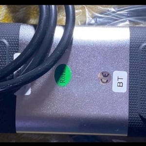 Könnte beinhalten: Ein rechteckiges Gerät mit silberner Mitte und schwarzen strukturierten Enden. Ein grüner "RoHS"-Aufkleber und ein weißer Aufkleber mit "BT" sind sichtbar. Schwarze Kabel sind über das Gerät geschlungen.