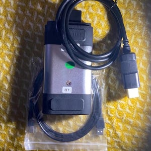 Könnte beinhalten: Silbernes und schwarzes Diagnosewerkzeug mit einem schwarzen Kabel und verschiedenen Anschlüssen. Das Gerät hat einen grünen Aufkleber und die Buchstaben "BT". Das Werkzeug befindet sich in einer transparenten Plastiktüte.