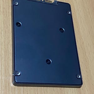 Op de afbeelding: Een zwarte solid-state drive (SSD) met een rechthoekige vorm. De SSD heeft een connector aan één uiteinde en verschillende schroeven om de behuizing vast te zetten. Het oppervlak is glad en reflecterend, met een donkerblauwe tint.