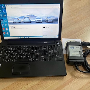Könnte beinhalten: Ein schwarzer Toshiba-Laptop, der die Benutzeroberfläche der Volvo VIDA-Fahrzeugdiagnosesoftware anzeigt, begleitet von einem Volvo DICE-Diagnosekommunikationstool mit angeschlossenem schwarzem Kabel und einer weißen Software-CD.