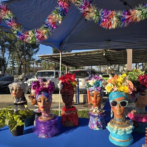May include: A table display of seven colorful mannequin heads decorated with flowers and fabric. Each head is wearing a different style of hat and has a unique design.