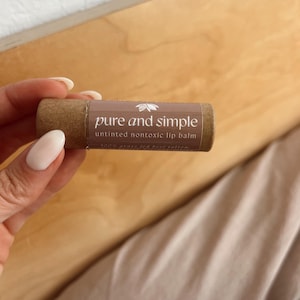 May include: A brown lip balm tube with the words "pure and simple" in white lettering. The text "untinted nontoxic lip balm" is below the main text. The lip balm is held in a person's hand.