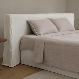 May include: A bed featuring a cream-coloured headboard and bed skirt. Two light brown pillows are on the bed, which is covered with a light brown blanket. The headboard has vertical seams with decorative stitching.