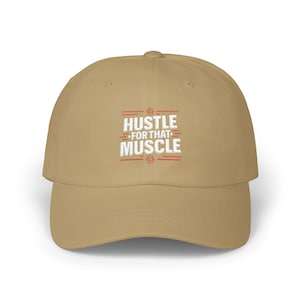May include: A tan baseball cap with the embroidered text "HUSTLE FOR THAT MUSCLE" in white and red. The cap has a curved brim and a six-panel design, suitable for casual wear.