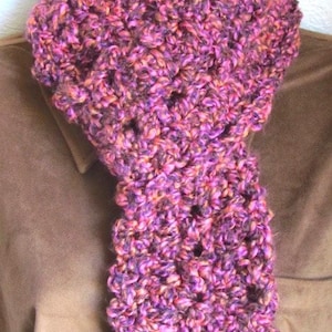 May include: A chunky, hand-knitted scarf in shades of purple, pink, and orange. The scarf is displayed on a mannequin, showcasing its textured, warm appearance. This accessory is perfect for adding a pop of color and warmth to any outfit.
