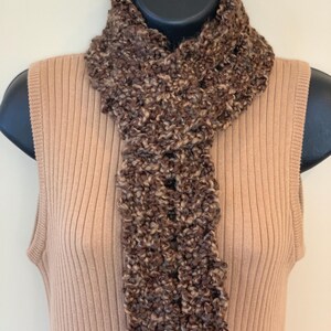 May include: A brown and tan textured scarf is draped over a mannequin. The scarf is made of a thick, looped yarn, creating a soft, cozy appearance. The color palette includes shades of brown and tan. The scarf is displayed on a tan ribbed tank top.