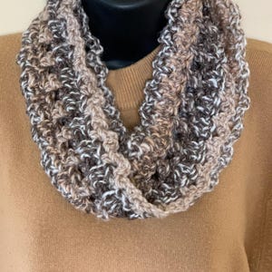 May include: A handmade infinity scarf in a neutral color palette of brown, beige, and white. The scarf is made of a chunky, textured yarn, and is displayed on a black mannequin head. The scarf is a cozy accessory.