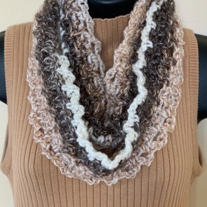 May include: A crocheted infinity scarf featuring a variegated yarn pattern in shades of cream, beige, and dark brown. The scarf has a textured, open-stitch design with a prominent white yarn border that creates a wavy, decorative edge throughout.