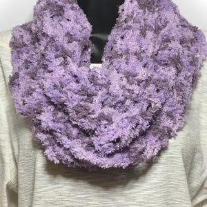 May include: A chunky infinity scarf made of fuzzy, textured yarn in varying shades of light and dark purple with subtle metallic silver flecks woven throughout the material, draped around a black display form over a white long-sleeve shirt.