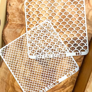 May include: Two white plastic stencils with a fish scale pattern. The stencils are labeled "SHY CAT STUDIO" and have the numbers "02-M" and "002".