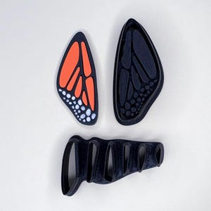 May include: Three-piece set of monarch butterfly-shaped cookie cutters. The set includes two wing-shaped cutters, one with orange and black details, and a body-shaped cutter. The cutters are black and orange.