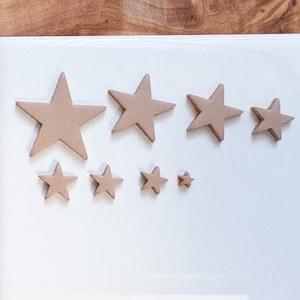 May include: A collection of tan-colored star-shaped decorations of varying sizes arranged on a white surface. The stars are made of a solid material and are arranged in a visually appealing pattern. The background is a light color.
