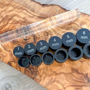 May include: A clear acrylic rolling pin and a set of black circular tools with measurements from 1 mm to 6 mm. The tools are arranged on a wooden surface with a natural grain pattern.