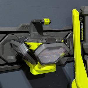May include: A wall-mounted RYOBI tool organizer with a gray and yellow color scheme. The organizer holds a work light and a stand-up light. The RYOBI logo is visible on the tools. The tools are mounted on a dark gray wall.
