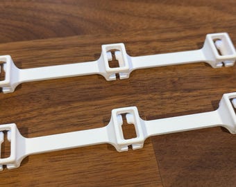 Op de afbeelding: Twee identieke witte kunststof verbindingsstukken, elk voorzien van een lange, platte centrale balk met drie rechthoekige openingen. Elke opening bevat een klein, flesvormig uitgesneden detail. De verbinders zijn stijve, lineaire onderdelen.
