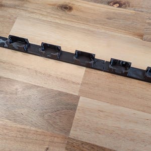 May include: A black plastic strip with multiple rectangular slots. The strip is lying on a wooden surface. The slots appear to be designed to hold or secure other objects. The overall design is simple and functional.