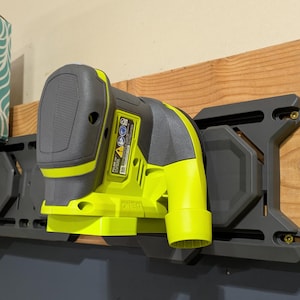 Può includere: Una levigatrice elettrica verde e nera è montata su un supporto da parete nero. La levigatrice ha una porta di aspirazione della polvere e un interruttore a grilletto. Il supporto ha una base in plastica nera e un inserto in plastica grigia.