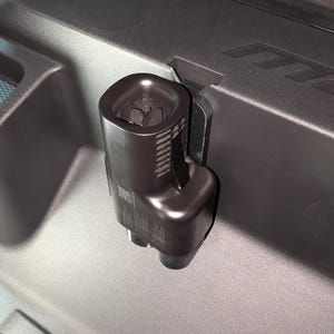 May include: A dark brown, cylindrical object with a textured surface and a small opening at the top. It is attached to a grey surface with a black bracket. The object appears to be a part of a larger mechanism or device.