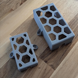 May include: Two gray, 3D-printed rectangular objects with a honeycomb pattern. The larger object has a series of hexagonal cutouts and mounting tabs. The smaller object has a similar design. Both objects are on a wooden surface.