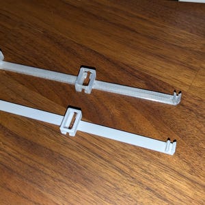 May include: Two white plastic cable ties with a rectangular design, lying on a wooden surface. Each tie has a central locking mechanism and two small prongs at the end. One tie is a lighter shade of white.