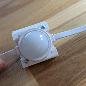May include: A white LED light module with a rounded, translucent lens, mounted on a white plastic frame. The module is secured with small screws and connected to a white wire. The item is on a wooden surface.