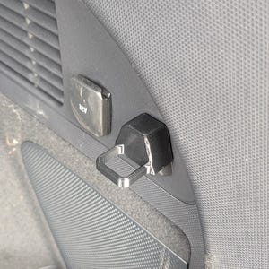 May include: A black plastic hook accessory mounted on a textured gray vehicle interior panel. The hook features a rectangular loop design and is positioned next to a 12V power outlet cover. The component is secured to the side wall of the car interior.
