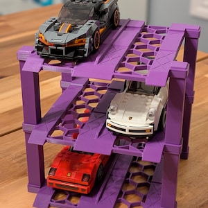 May include: A three-tiered purple plastic car parking garage with three toy cars on it. The top car is black and orange, the middle car is white, and the bottom car is red.