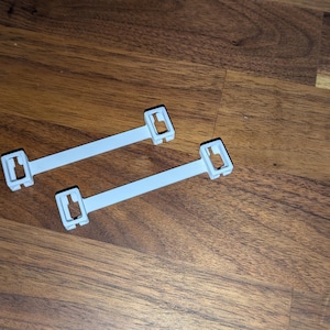 May include: Two light grey plastic cable ties with rectangular ends and a central bar. The ties are lying on a wooden surface with a natural wood grain pattern. The ties are designed to secure cables.