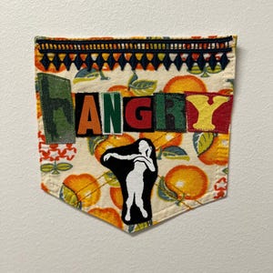 May include: A decorative pocket patch with the word "ANGRY" in colorful block letters. The patch features a pattern of oranges and leaves, with a black and white silhouette of a person dancing. The top edge has a decorative trim.