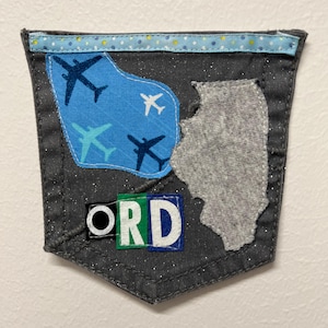 May include: A gray fabric pocket with a blue patterned border. The pocket features a blue patch with airplane designs, a gray Illinois state shape, and the letters "ORD" in white, green, and blue.