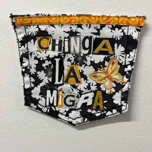 May include: A fabric pocket with a black and white floral pattern. The pocket features the words "CHINGA LA MIGA" in a mix of block letters and a butterfly applique. The top edge has a decorative yellow and orange trim.