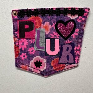 May include: A decorative denim pocket with a floral pattern in shades of pink and purple. The word "PLUR" is spelled out with colorful fabric letters. A glittery pink heart and black lace trim add detail.