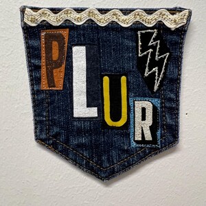 May include: A denim pocket with the letters "PLUR" appliqued in various colors. The pocket is trimmed with a gold and white decorative trim. A black lightning bolt patch is also present.
