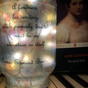 May include: A glass jar with a quote about reading, "A fondness for reading, which properly directed must be an education in itself." The jar is decorated with a floral pattern and has a string of lights inside. The jar is sitting on a keyboard.