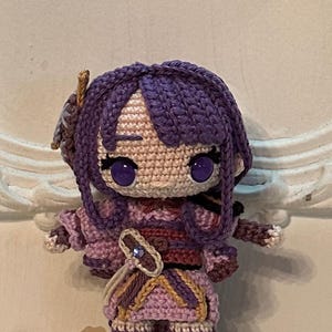 May include: Crocheted doll with purple hair, pink and brown clothing, and large purple eyes.