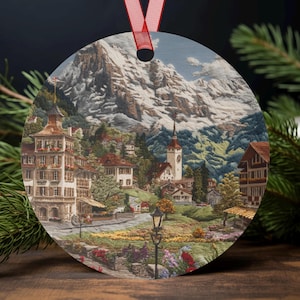 Puede incluir: Adorno navideño redondo que representa un paisaje detallado de un pueblo de montaña con picos nevados. El adorno tiene una cinta roja para colgar. La imagen incluye edificios, árboles y una iglesia.