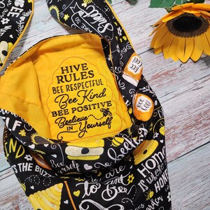 May include: A yellow fabric tote bag with black and yellow bee-themed print. The bag has a pocket on the front with the text "Hive Rules Bee Respectful Bee Kind Bee Positive Beelieve in Yourself".