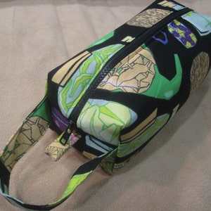 May include: A black zippered travel bag with a green, yellow, and brown patterned design. The bag has a long strap and a zipper closure.