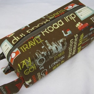 May include: Brown travel-themed zippered pouch with a handle. The pouch has a zipper closure and is printed with the words "TRAVEL Road Trip" and various city names, including Paris, London, and Berlin.
