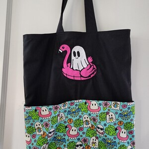 May include: Black tote bag with a colorful bottom panel. The top of the bag features an embroidered ghost floating on a pink flamingo float. The bottom panel has a repeating pattern of ghosts, flamingos, and tropical leaves.